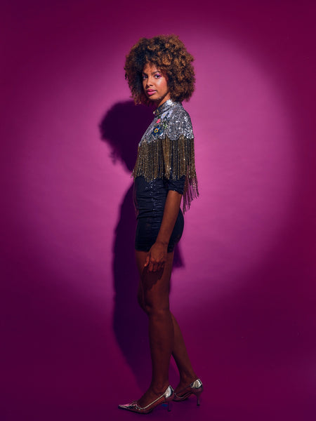 Person wearing a sequinned Cape with fringing and black shorts against a pink background