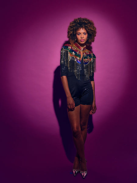 Woman wearing a colorful top with fringe and black shorts against a purple background