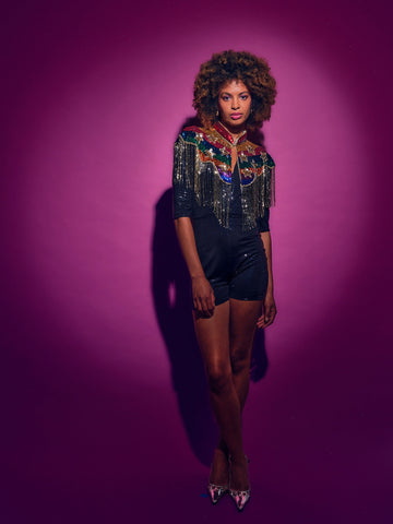 Woman wearing a colorful top with fringe and black shorts against a purple background