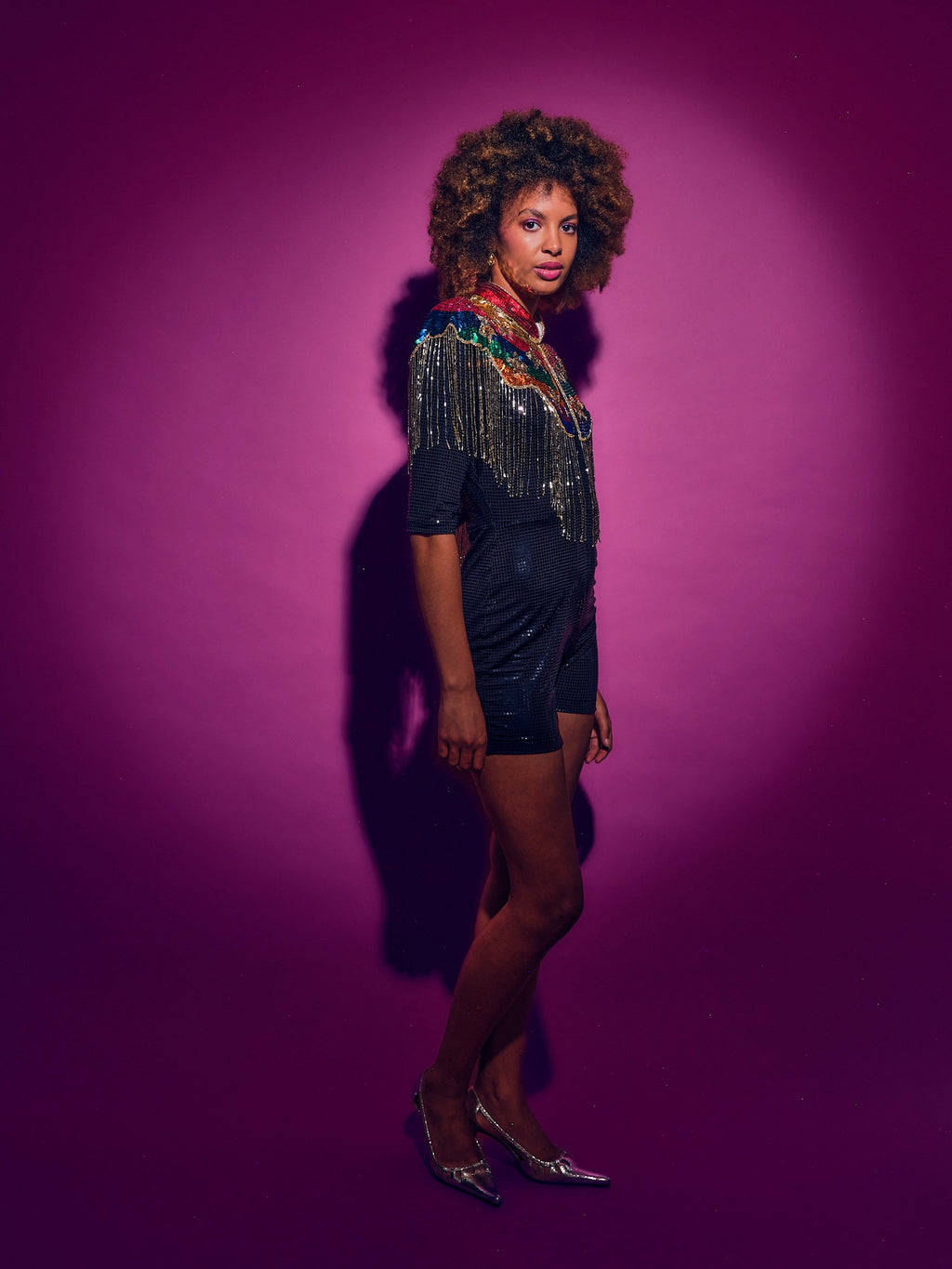 Woman in a black dress with sequins and a colorful rainbow cape against a pink background