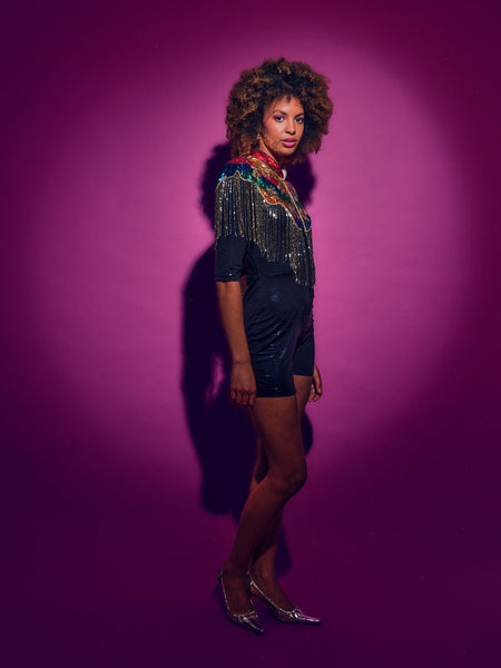 Woman in a black dress with sequins and a colorful rainbow cape against a pink background