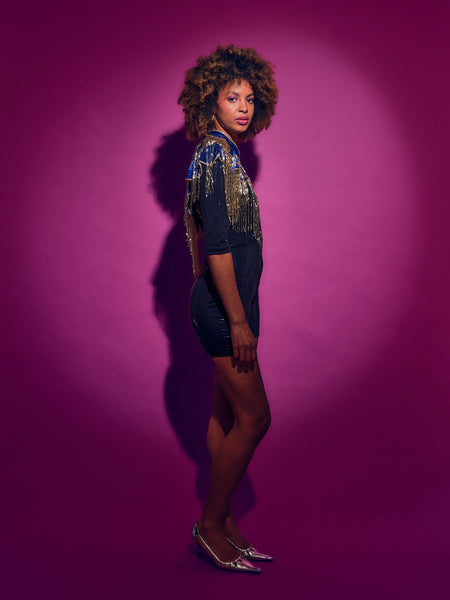 Person wearing a Blue and Gold sequinned Cape with fringing and black shorts against a pink background
