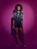 Person wearing a Green and silver sequinned Cape with fringing and black shorts against a pink background