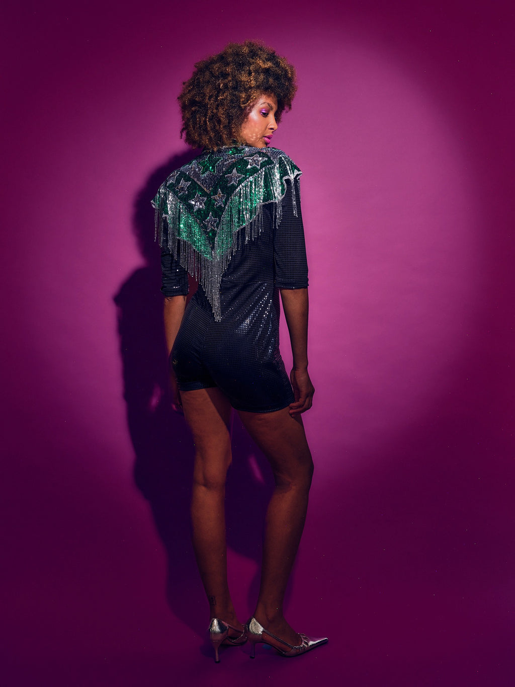 Person wearing a Green and silver sequinned Cape with fringing and black shorts against a pink background