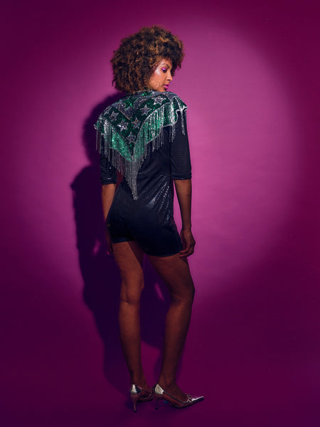 Person wearing a Green and silver sequinned Cape with fringing and black shorts against a pink background