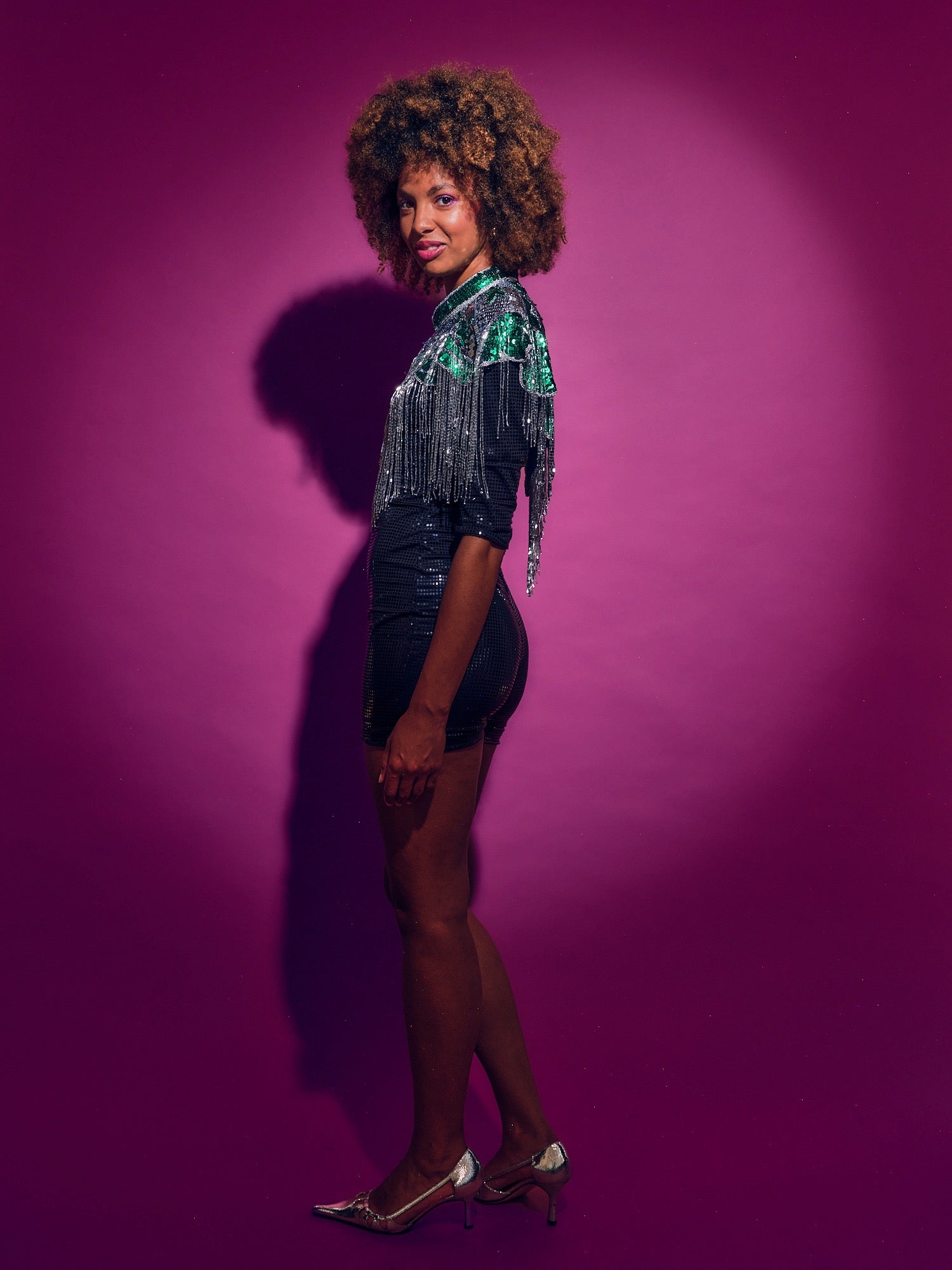 Person wearing a Green and silver sequinned Cape with fringing and black shorts against a pink background
