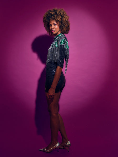 Person wearing a Green and silver sequinned Cape with fringing and black shorts against a pink background