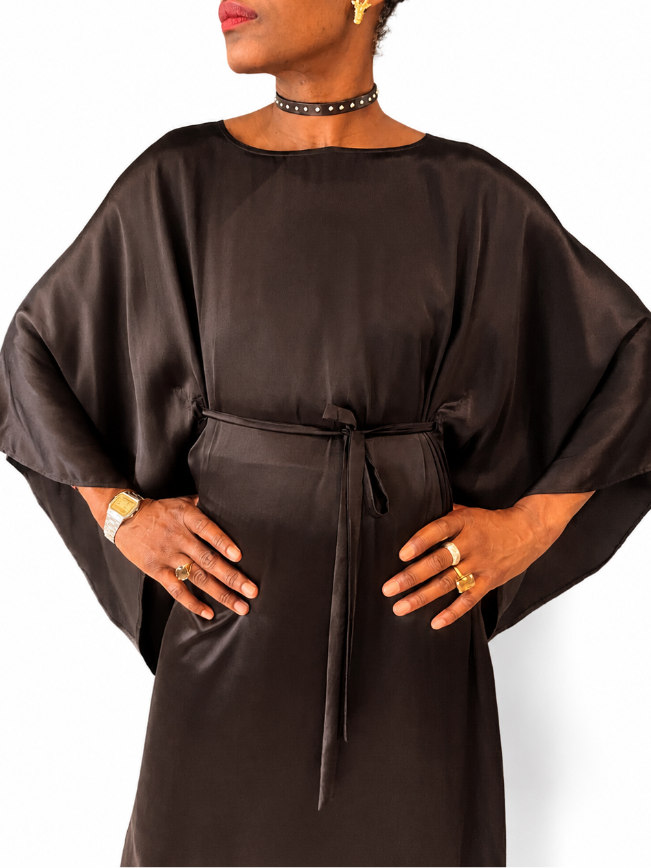 A woman modeling a dark brown satin-like dress with wide draped sleeves and a self-tie waist belt, photographed against a plain white background.