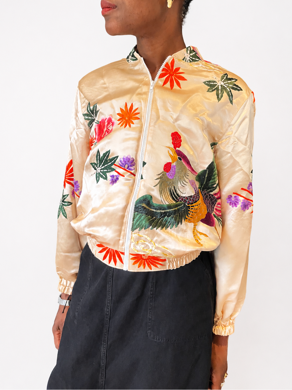 A model wearing a shiny beige satin bomber jacket with colorful embroidered tropical flowers and a rooster, paired with a dark gray skirt, against a plain white background.