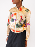 A model wearing a shiny beige satin bomber jacket with colorful embroidered tropical flowers and a rooster, paired with a dark gray skirt, against a plain white background.