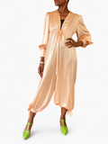 A woman modeling a peach satin jumpsuit with a tie waist and long sleeves, paired with bright green heels, against a plain white studio background.