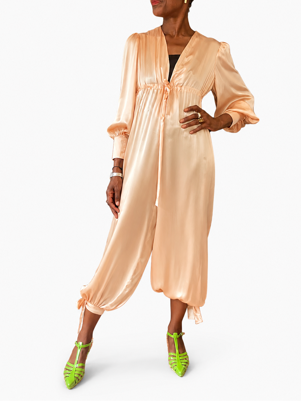 A woman modeling a peach satin jumpsuit with a tie waist and long sleeves, paired with bright green heels, against a plain white studio background.