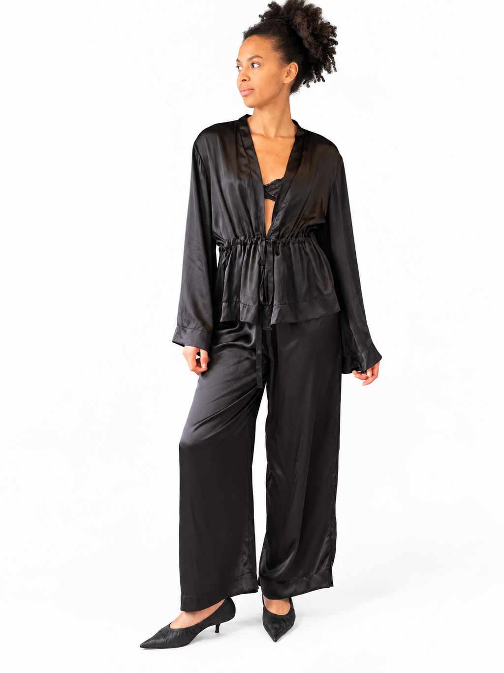 A woman modeling a black satin two-piece loungewear set consisting of a long-sleeve V-neck top and matching wide-leg pants, paired with black pointed-toe heels against a plain white background.