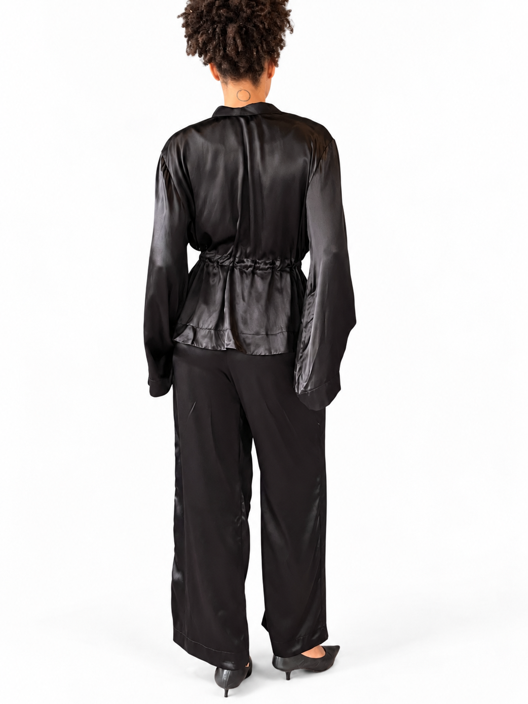 A model shown from the back wearing a black satin long-sleeve top with a gathered peplum waist and black wide-leg trousers, photographed against a plain white studio background.