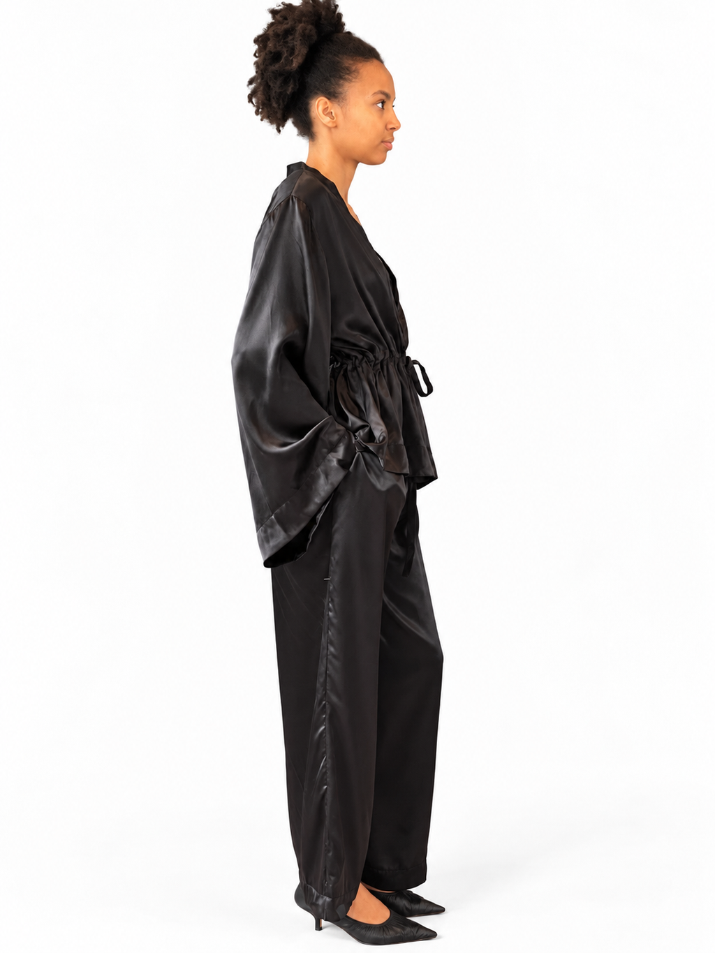 A woman shown in side profile wearing a black satin-like robe with wide draped sleeves and matching wide-leg pants, photographed against a plain white studio background.